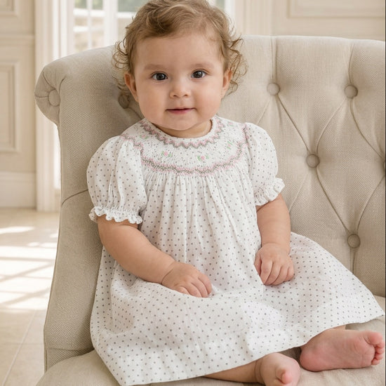 Baby girl wearing a Carriage Boutique off-white smocked dress with black polka dots and floral embroidery