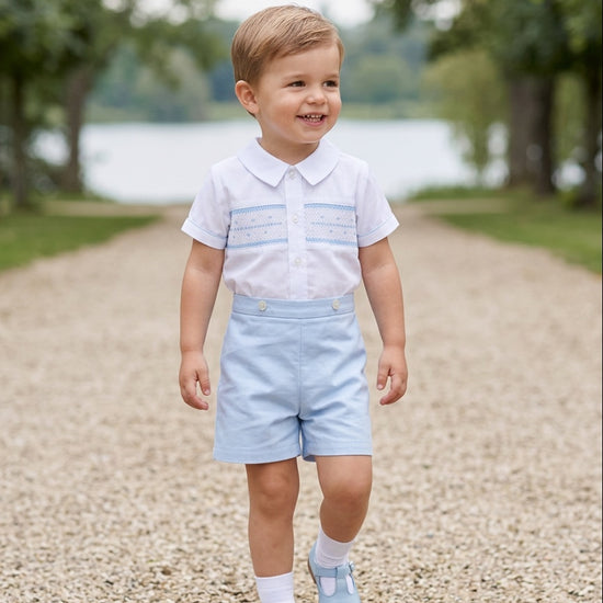 Carriage Boutique toddler boy wearing a white smocked shirt and light blue shorts bobby suit on a gravel path