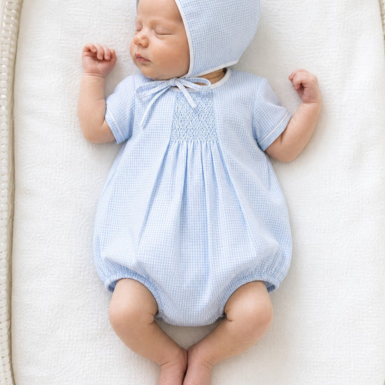 Carriage Boutique light blue gingham smocked bubble and bonnet set with white piping. Baby lying down.