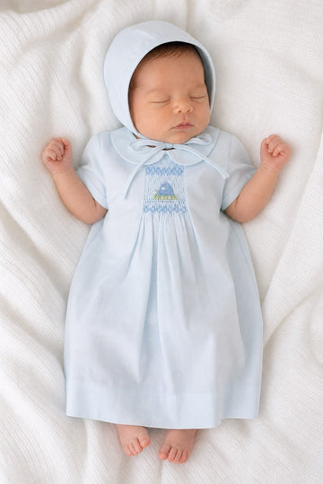 Newborn baby wearing a Carriage Boutique baby blue smocked heirloom dress and matching bonnet on a white blanket.