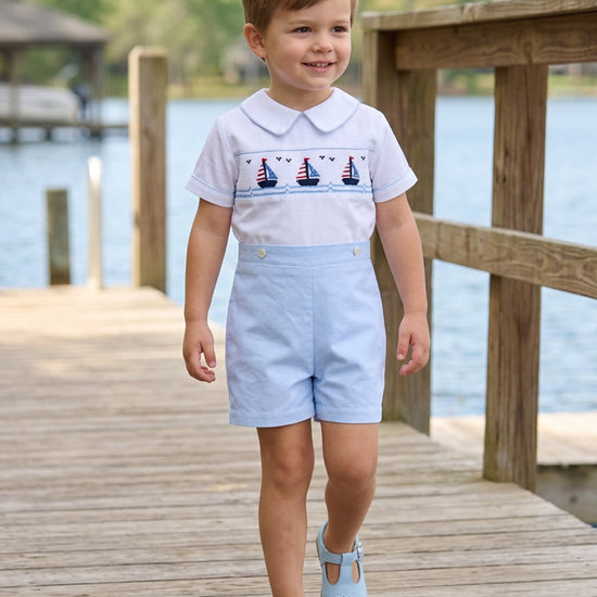 Carriage Boutique toddler boy wearing a white smocked sailboat bobby suit with light blue shorts on a wooden dock.