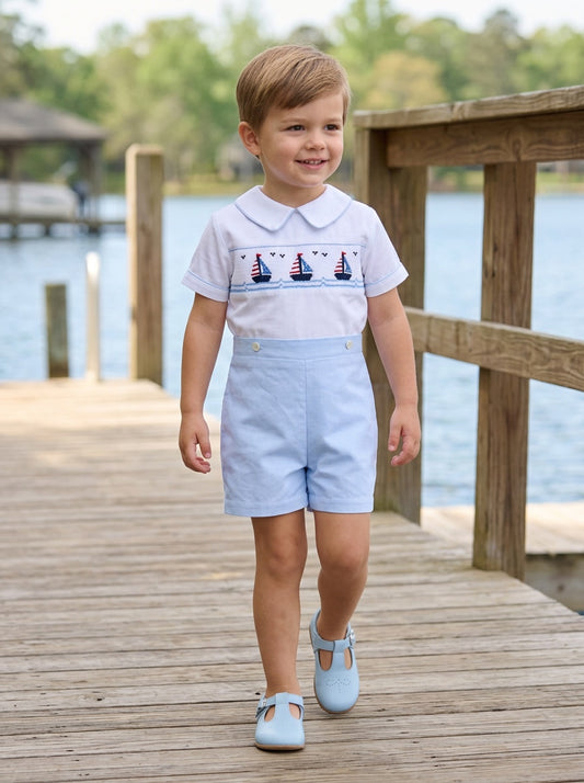 Carriage Boutique toddler boy wearing a white smocked sailboat bobby suit with light blue shorts on a wooden dock.