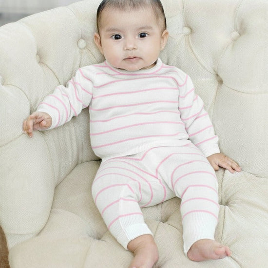 Baby wearing a Carriage Boutique white and blush pink striped cotton long-sleeve legging set while sitting on a chair.