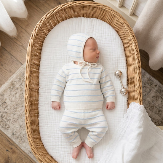 Carriage Boutique baby sleeping in a wicker basket wearing a white and sky-blue striped knit legging set with a hat.