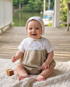 A happy baby boy wearing a Carriage Boutique tan and white bubble romper with a matching bonnet on a porch.
