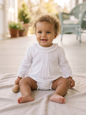 Carriage Boutique baby girl white long sleeve bubble romper with smocked collar sitting on a soft blanket