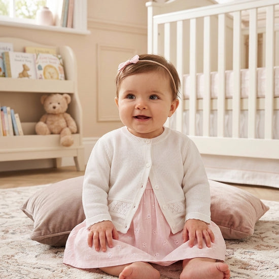 Carriage Boutique baby girl wearing a white knit cardigan over a pink dress in a nursery setting.