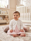 Carriage Boutique baby girl wearing a white knit cardigan over a pink dress in a nursery setting.