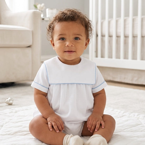 Carriage Boutique baby boy wearing a white cotton romper with blue trim and a classic collar, sitting in a nursery.