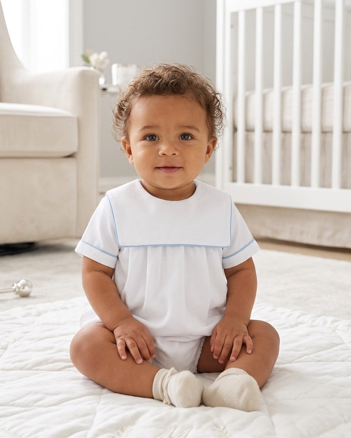 Carriage Boutique baby boy wearing a white cotton romper with blue trim and a classic collar, sitting in a nursery.