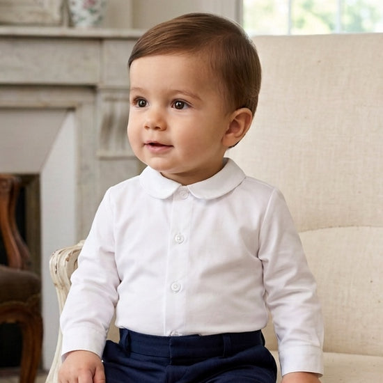 Carriage Boutique baby boy wearing a white long sleeve button-down shirt with a rounded collar sitting on a chair