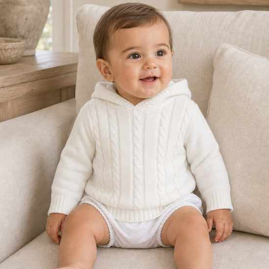 Carriage Boutique white cotton cable knit hooded baby sweater worn by a smiling baby sitting on a beige sofa.