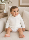 Carriage Boutique white cotton cable knit hooded baby sweater worn by a smiling baby sitting on a beige sofa.