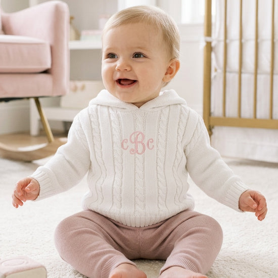 Carriage Boutique white cable knit hooded baby sweater with pink monogram, worn by a smiling baby in a nursery.