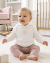 Carriage Boutique white cable knit hooded baby sweater with pink monogram, worn by a smiling baby in a nursery.