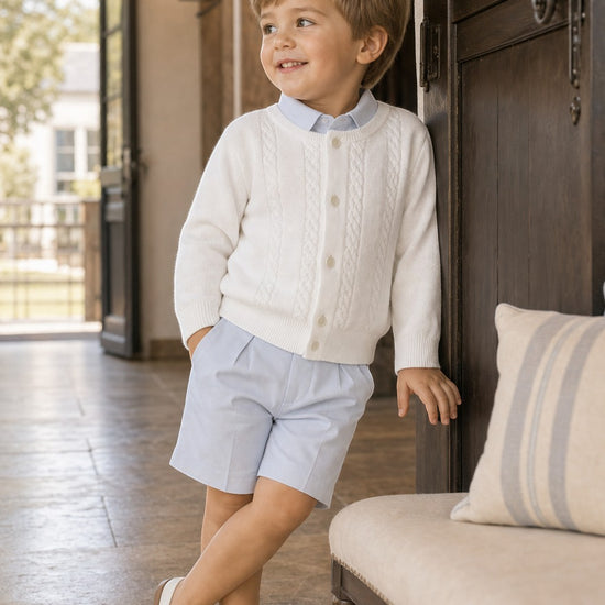 Carriage Boutique white cable-knit cotton cardigan worn by a young boy with a light blue shirt and shorts