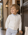 Carriage Boutique white cable-knit cotton cardigan on a young boy wearing a light blue shirt and trousers