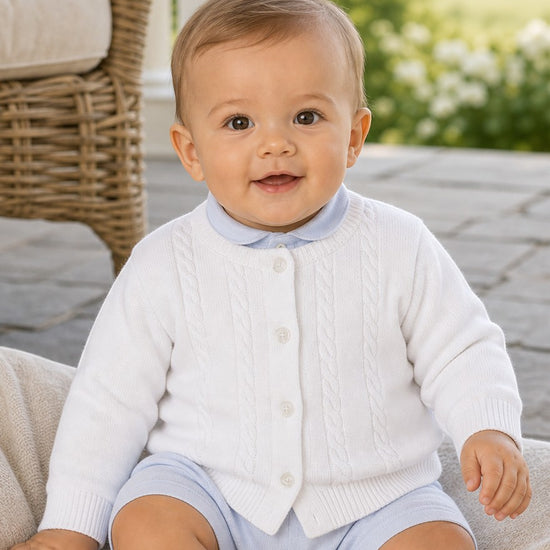 Carriage Boutique baby boy wearing a white cable-knit cotton cardigan with a button-front over a light blue outfit.