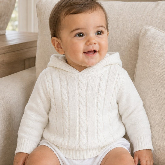 Carriage Boutique white cotton cable knit hooded baby sweater worn by a smiling baby sitting on a chair