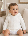 Carriage Boutique white cotton cable knit hooded baby sweater worn by a smiling baby sitting on a chair