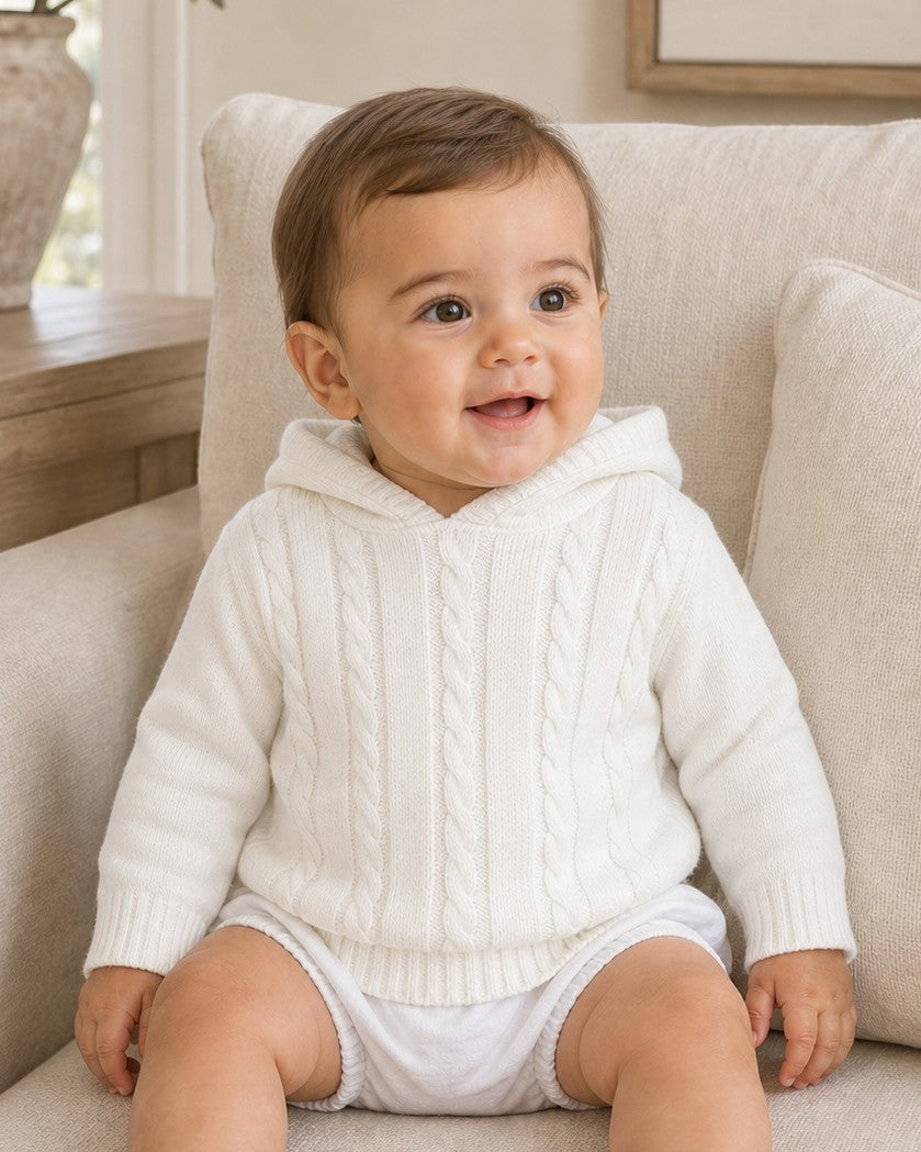 Carriage Boutique white cotton cable knit hooded baby sweater worn by a smiling baby sitting on a chair