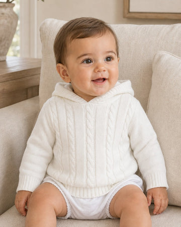 Carriage Boutique white cable knit hooded baby sweater made of soft cotton worn by a smiling baby