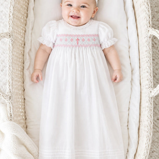 Carriage Boutique baby girl christening gown in white with pink hand-smocked yoke, embroidered cross, and ruffles.