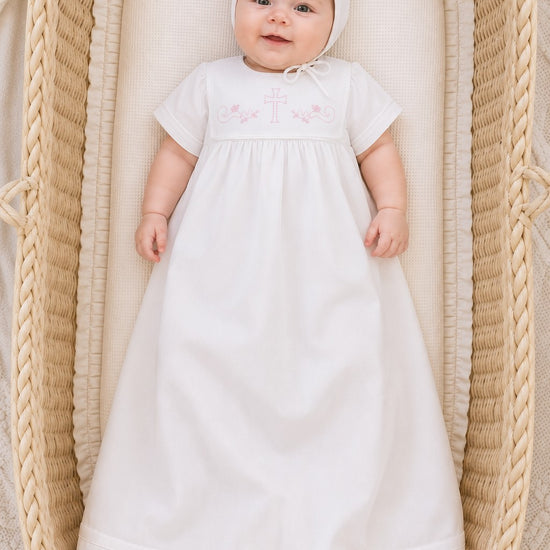 Carriage Boutique white baby christening gown with matching bonnet and pink cross embroidery in a wicker bassinet
