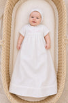 Carriage Boutique white baby christening gown with matching bonnet and pink cross embroidery in a wicker bassinet