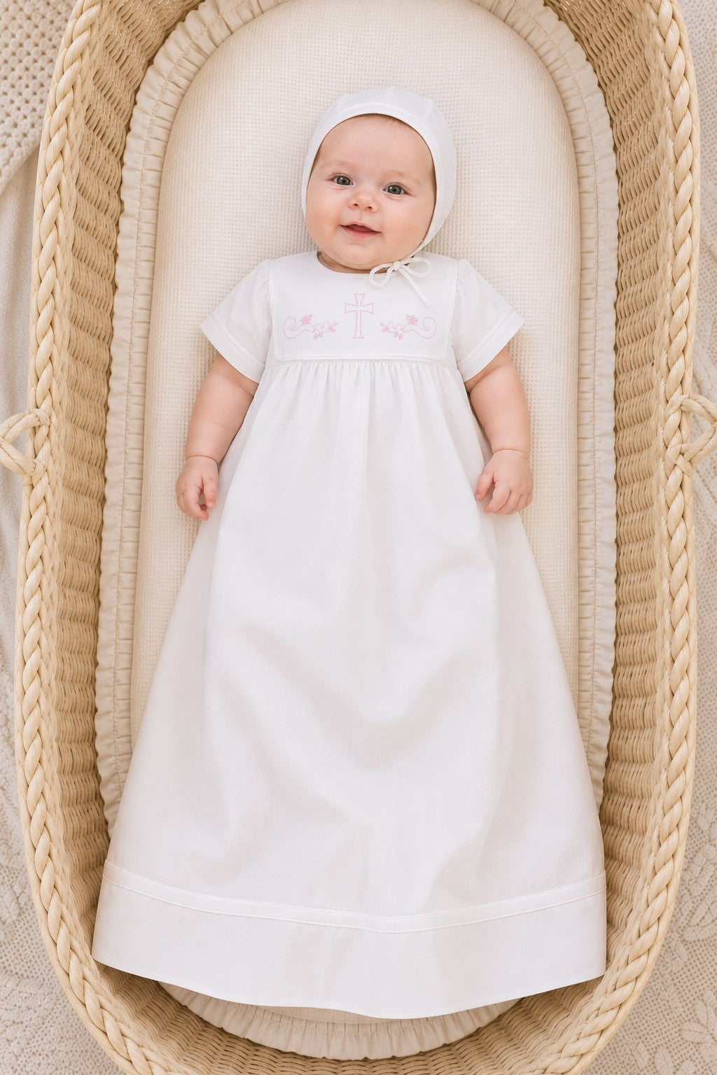 Carriage Boutique white baby christening gown with matching bonnet and pink cross embroidery in a wicker bassinet