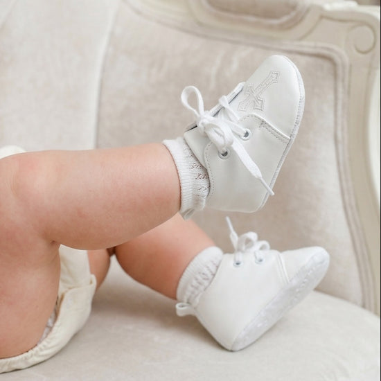 Carriage Boutique white baby boy christening shoes with cross embroidery and lace-up closure on a soft chair