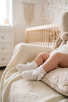 Carriage Boutique white baby girl christening socks with delicate lace trim and a satin bow detail on a baby's feet.