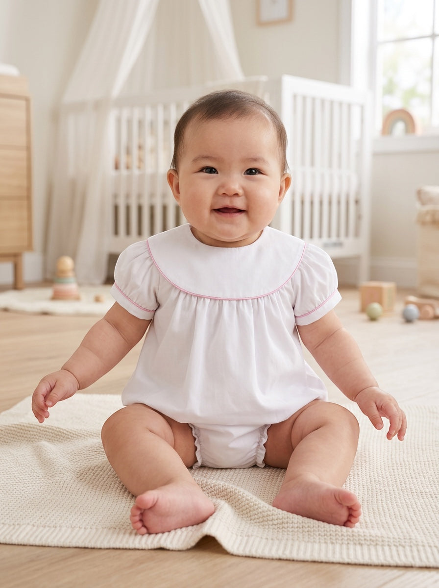 Carriage Boutique white cotton baby girl bubble romper with flutter sleeves and pink picot trim in a nursery setting