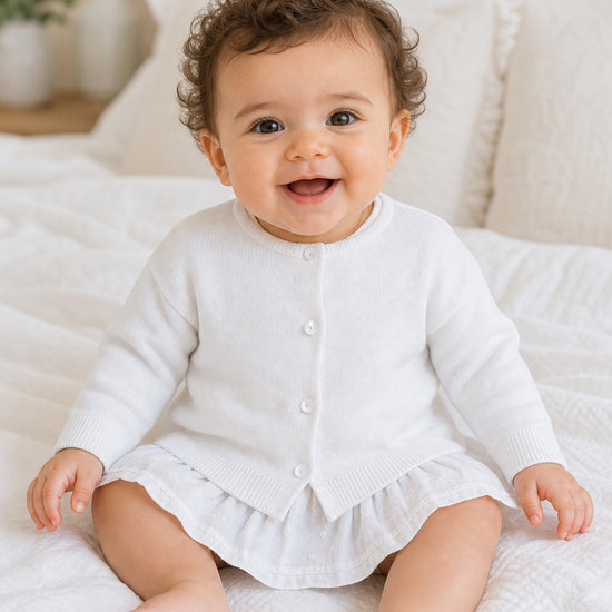 Carriage Boutique baby girl wearing a white cotton cashmere cardigan with ribbed hems and button front on a bed