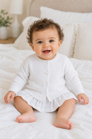 Carriage Boutique baby girl wearing a white cotton cashmere cardigan with ribbed hems and button front on a bed
