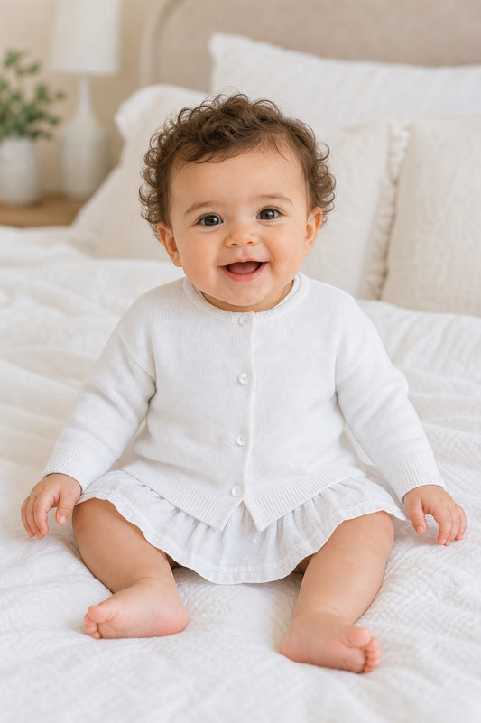 Carriage Boutique baby girl wearing a white cotton cashmere cardigan with ribbed hems and button front on a bed
