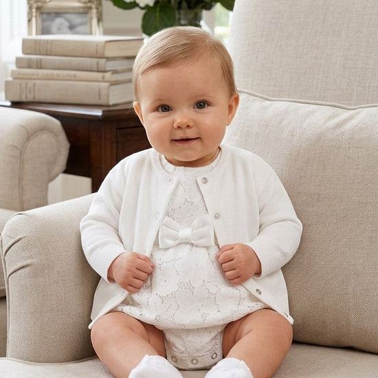 Carriage Boutique white cotton cashmere baby girl cardigan worn over a matching lace romper while sitting on a chair