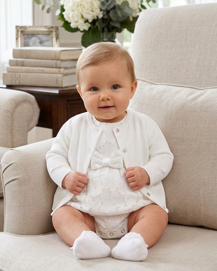 Carriage Boutique white cotton cashmere baby girl cardigan worn over a matching lace romper while sitting on a chair