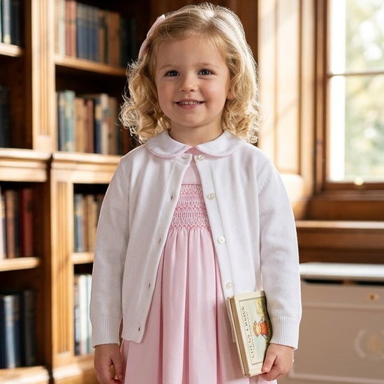 Carriage Boutique white cotton cashmere baby girl cardigan worn over a pink smocked dress in a library setting.