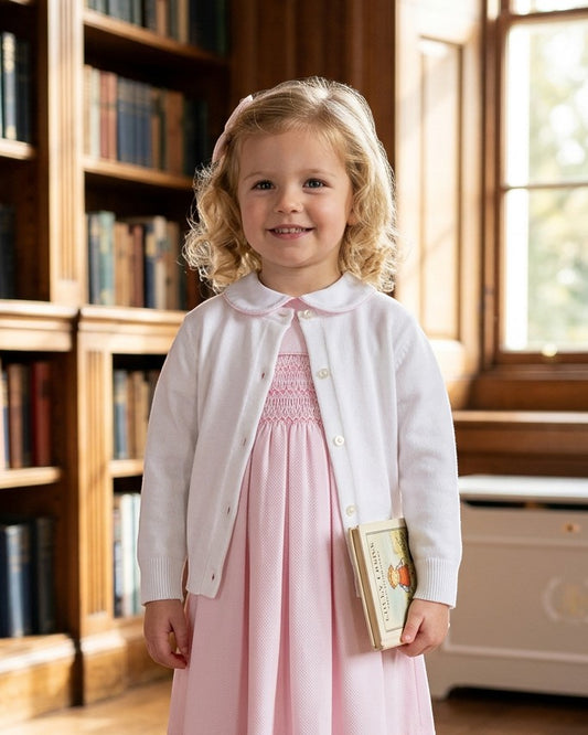 Carriage Boutique white cotton cashmere baby girl cardigan worn over a pink smocked dress in a library setting.