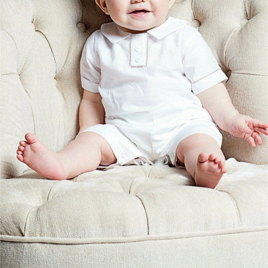 Carriage Boutique baby boy wearing a white cotton christening shortall with hand embroidery sitting on a tufted chair.