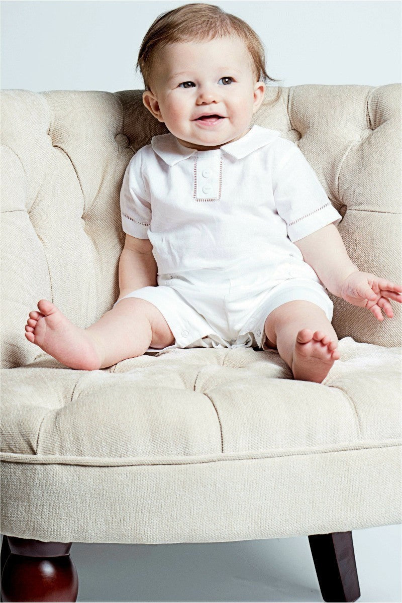 Carriage Boutique baby boy wearing a white cotton christening shortall with hand embroidery sitting on a tufted chair.