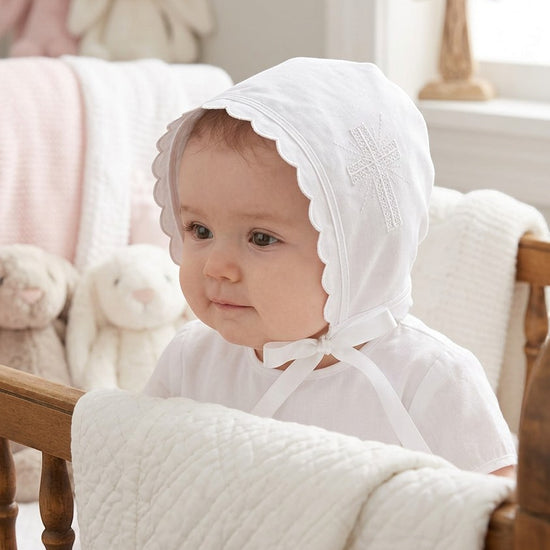 Carriage Boutique white cotton christening bonnet with embroidered cross, scalloped edges, and satin ribbon ties.