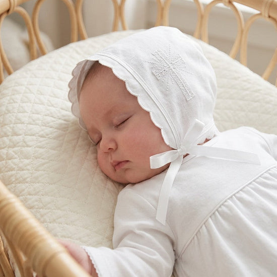 Carriage Boutique white cotton christening bonnet with embroidered cross, scalloped edge, and satin ribbon tie on baby