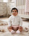 Carriage Boutique white cotton hooded baby sweater worn by a smiling baby girl sitting on a nursery rug.