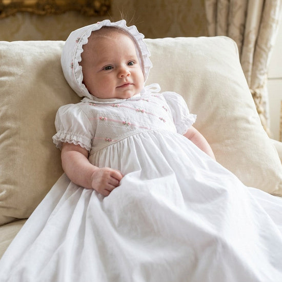 Carriage Boutique white infant gown with pink ribbon and floral embroidery, featuring a matching ruffled bonnet.