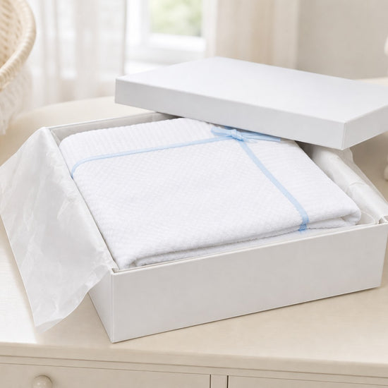 Carriage Boutique white knit baby blanket with blue satin ribbon and bow in a gift box on a nursery table.