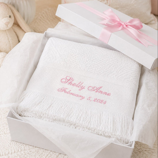 Carriage Boutique white knit baby blanket with pink personalized embroidery and fringe in a gift box with pink ribbon.
