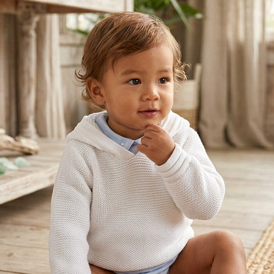 Carriage Boutique white knit baby boy hoodie with hood, worn by a toddler in a bright, rustic indoor setting.