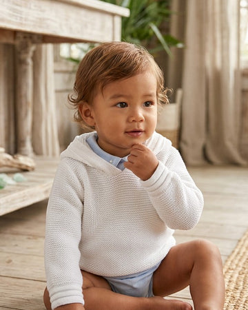 Carriage Boutique white knit baby boy hoodie with hood, worn by a toddler in a bright, rustic indoor setting.